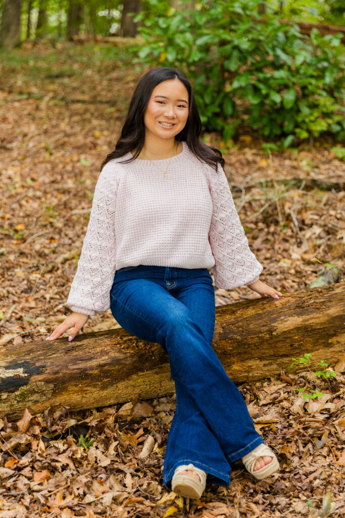 High school senior Mia sitting on a tree log in Brookhaven wearing jeans and a pink sweater.