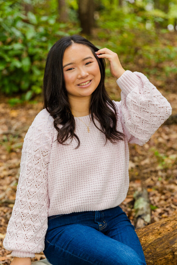 Mia sitting cross-legged on a tree log, touching her hair during her senior session in Brookhaven.
