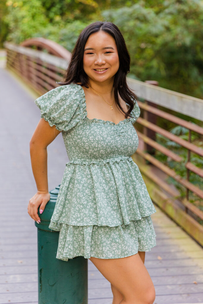 Mia leaning against a pole at the entrance of a Brookhaven park bridge wearing green and white jumper dress.