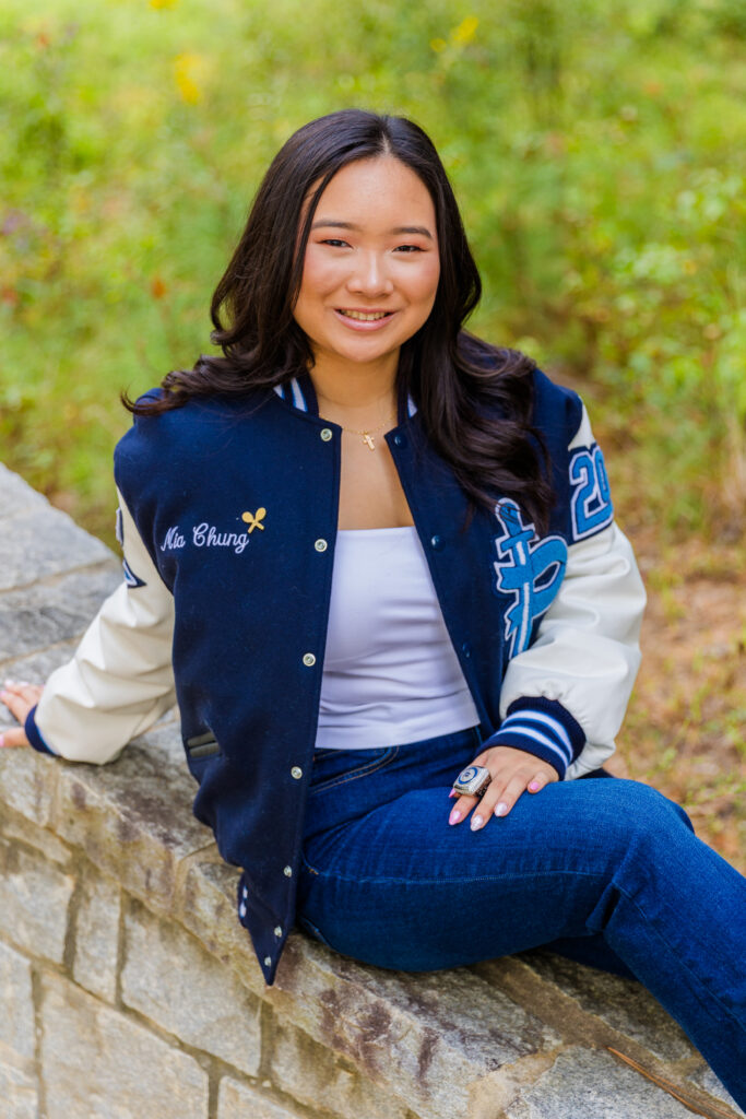 Senior portrait of Mia sitting on a wall in Brookhaven park wearing jeans, white top, leather jacket, and school ring.