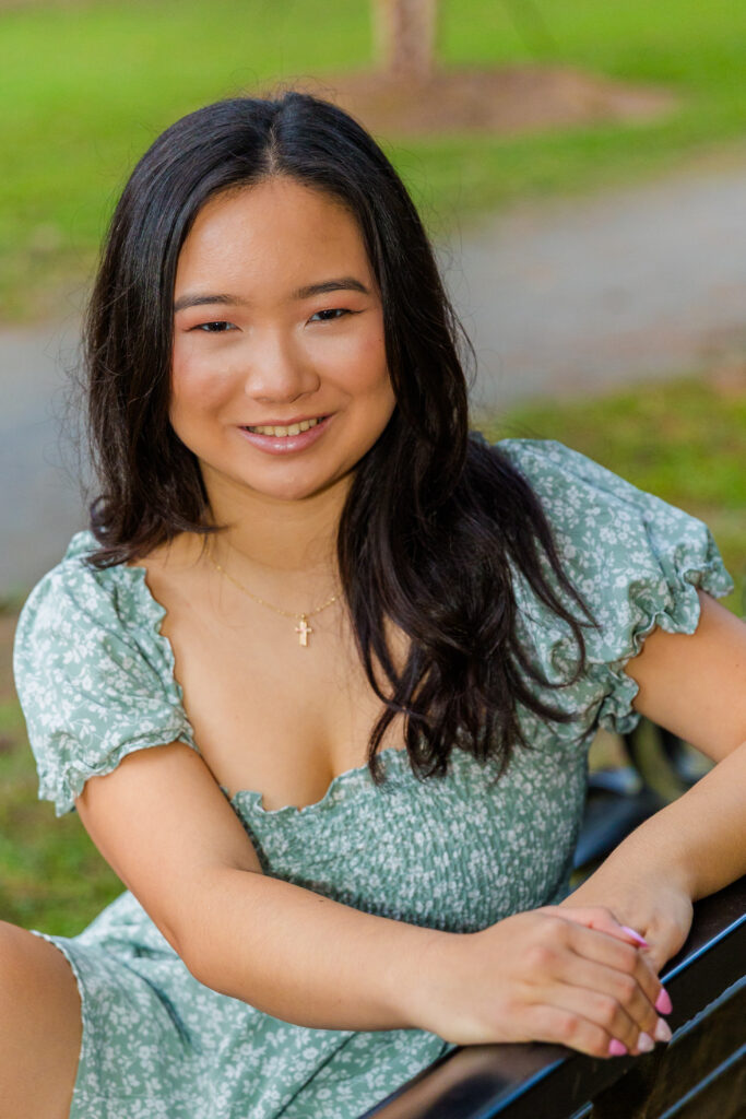 Pace Academy senior sitting on swing bench in Brookhaven park wearing green and white jumper dress, hands together.