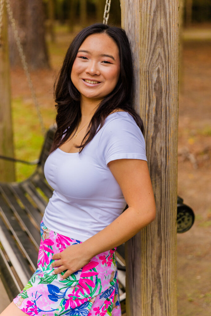 Mia leaning against a wooden swing pole in Brookhaven park, one leg up, wearing colorful skirt and white top.