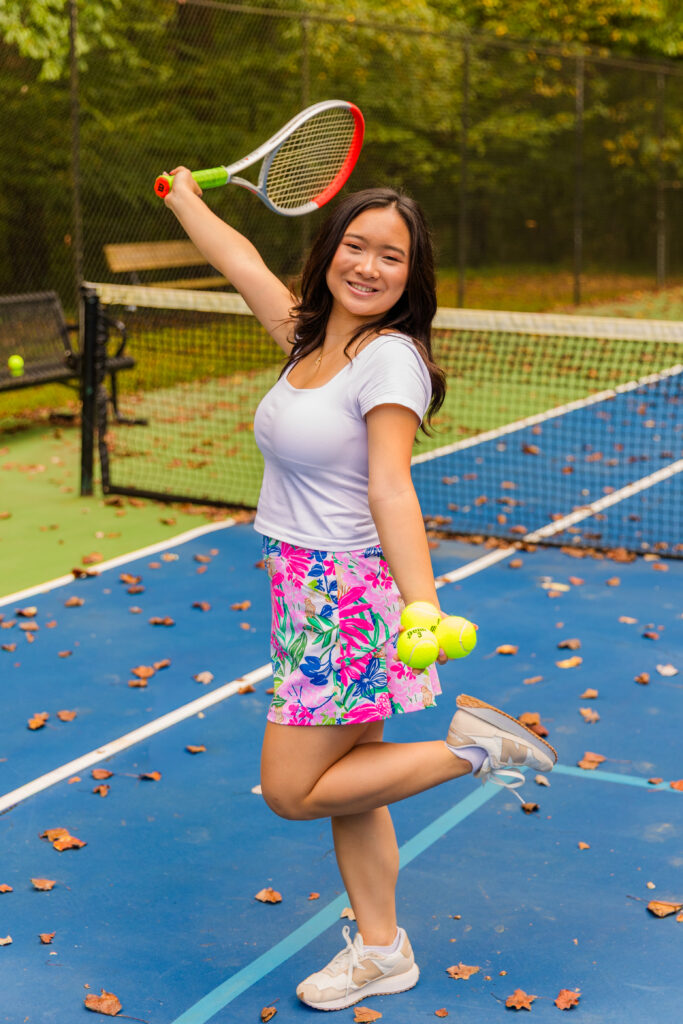 High school senior holding tennis racket and balls on court, one foot up, smiling in Atlanta.