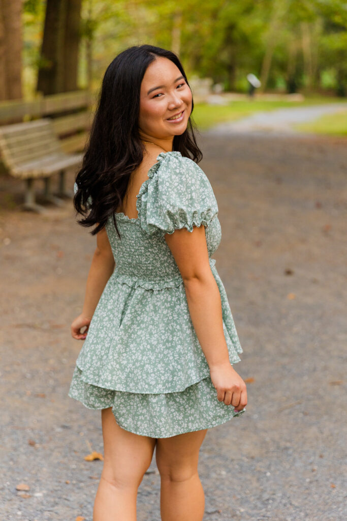 High school senior walking away from camera looking over shoulder in Brookhaven park in green and white dress.