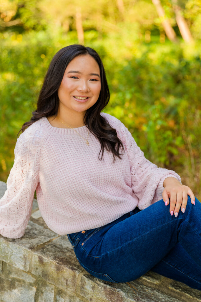 Senior portrait of Mia sitting on a stone wall in Brookhaven wearing a pink sweater and jeans.