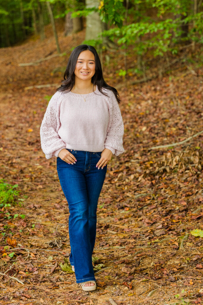 Pace Academy senior Mia walking in the woods wearing blue jeans and a light pink sweater.