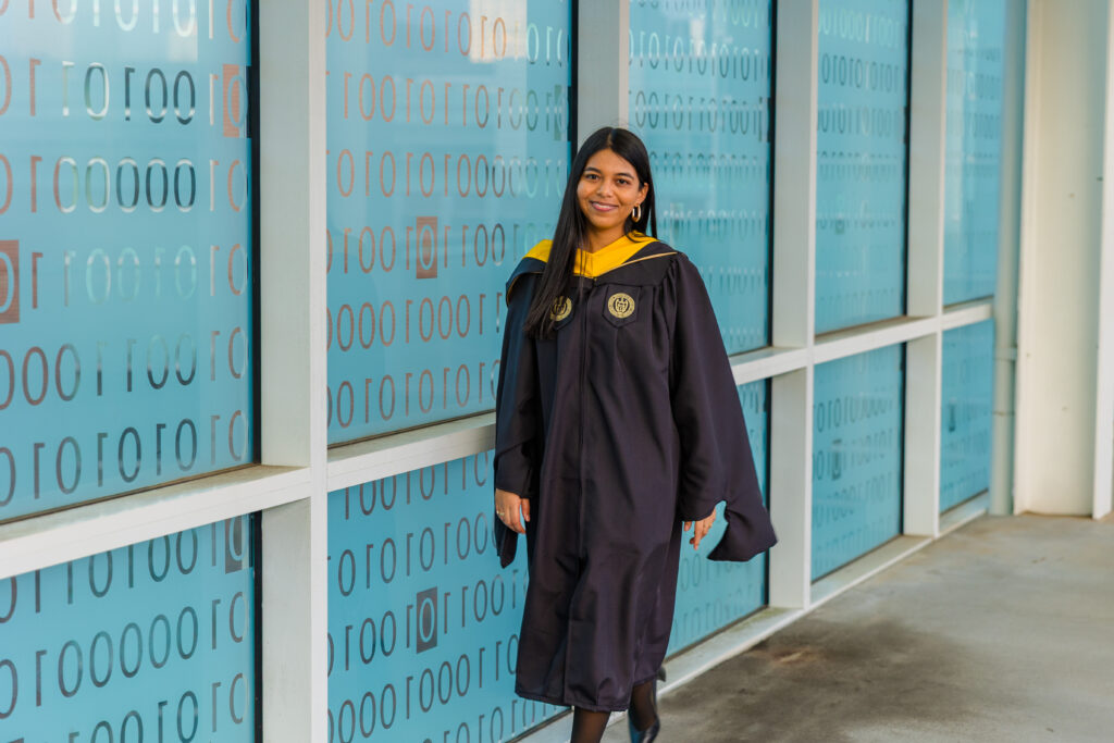 Pallovi walking alongside the transparent binary code wall on Georgia Tech campus in graduation attire