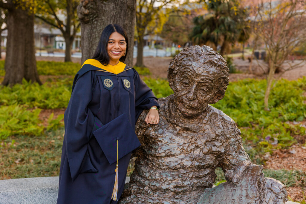 Wide golden hour graduation photo of Pallovi walking toward the camera with a statue behind her on campus
