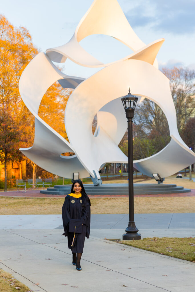 Wide golden hour graduation photo of Pallovi walking toward the camera with a statue behind her on campus