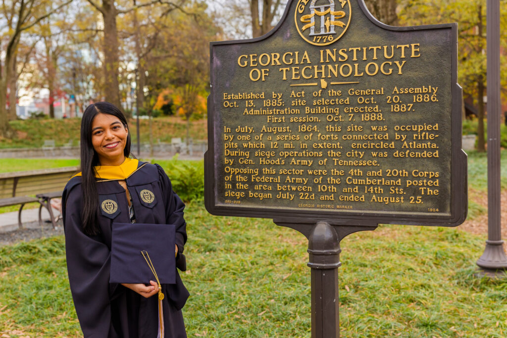 Pallovi standing in front of the Georgia Tech campus sign wearing her graduation cap and gown in Atlanta
