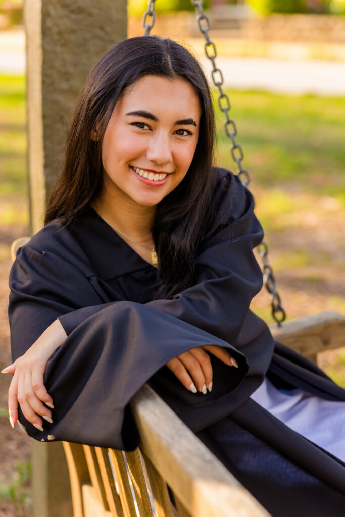 Senior on swing bench looking over shoulder wearing graduation gown in Atlanta park
