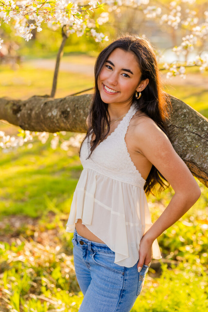 Senior in jeans and white sleeveless top leaning against blooming tree in Piedmont Park Atlanta
