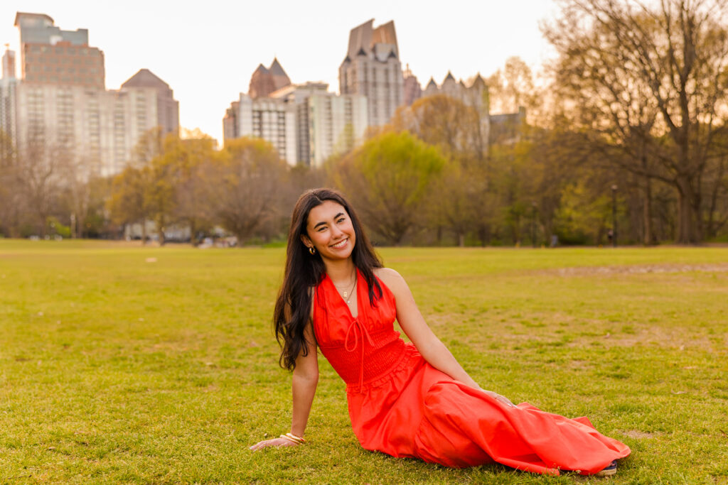 Wide shot of senior in orange dress sitting on grass with Atlanta skyline