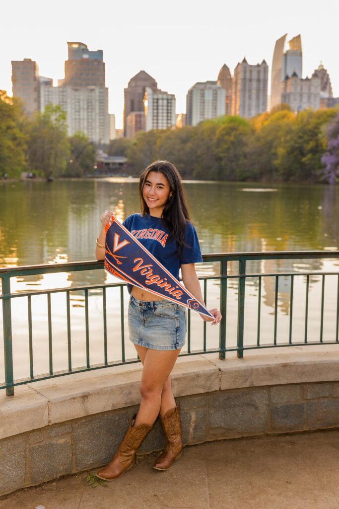 Senior in UVA shirt, denim skirt and cowboy boots holding banner with Atlanta skyline