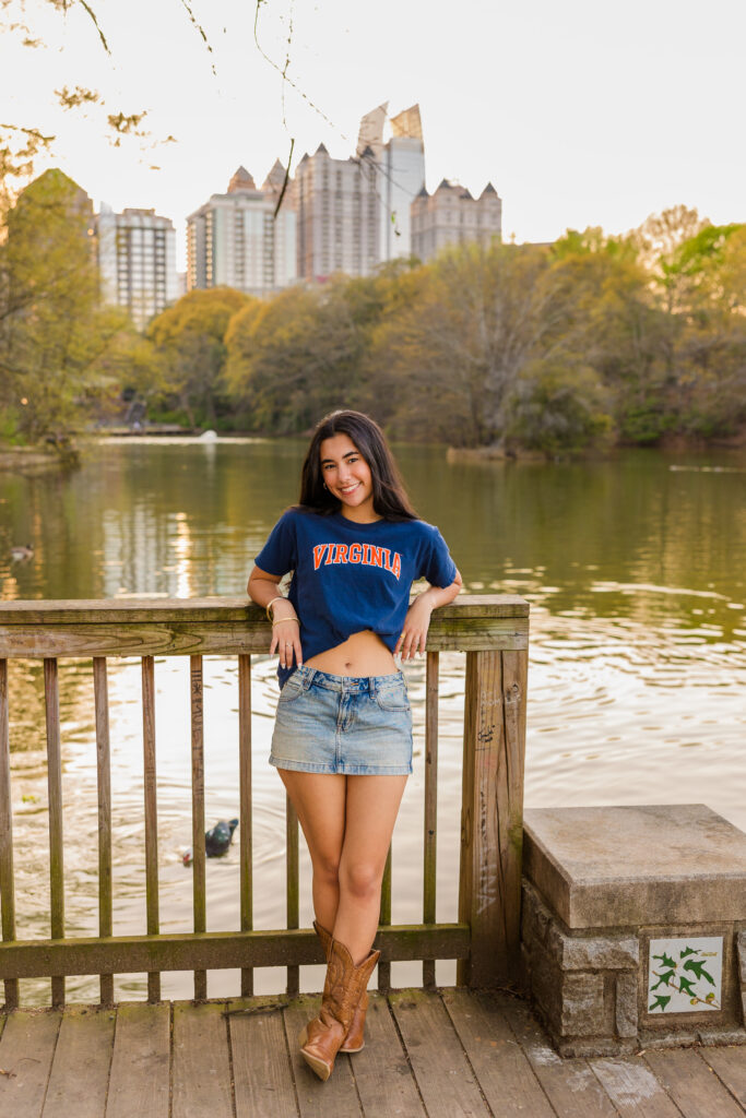 Senior leaning on wooden fence with Piedmont Park pond and Atlanta skyline