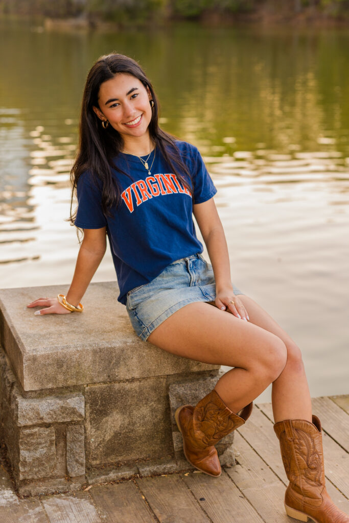Senior sitting on rock by pond wearing UVA shirt and denim skirt
