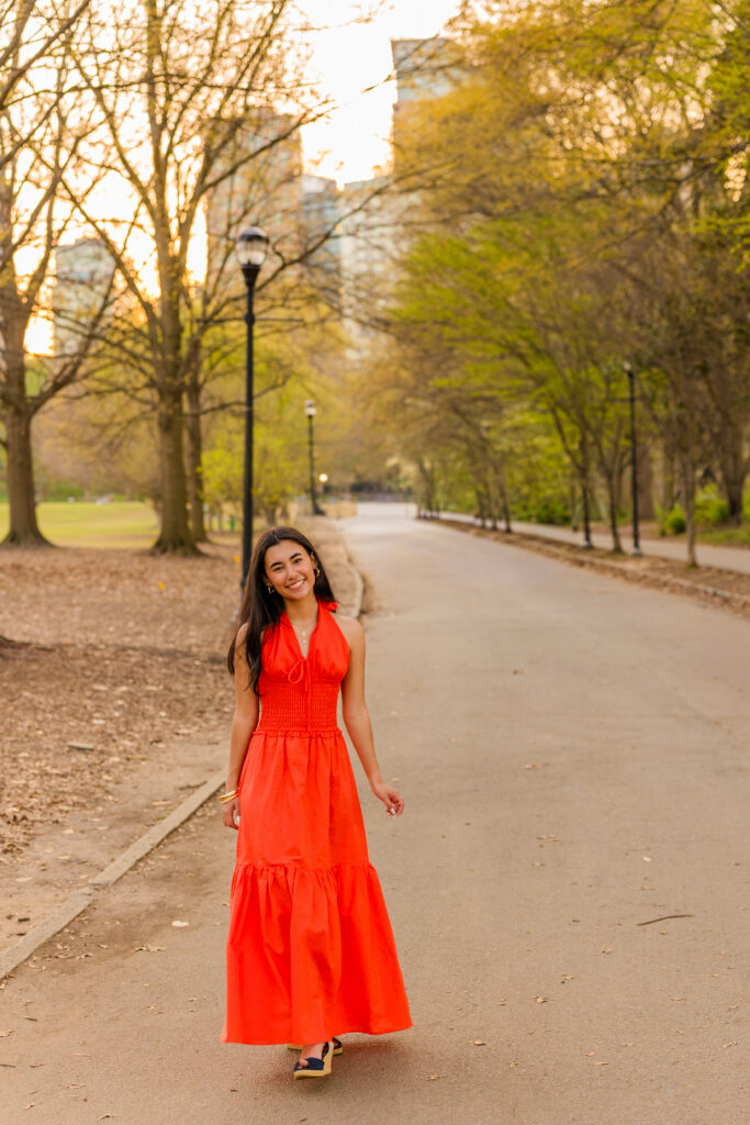Senior walking in orange dress with Atlanta skyline in Piedmont Park