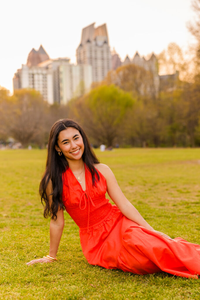 Senior sitting on grass in orange dress with Atlanta skyline