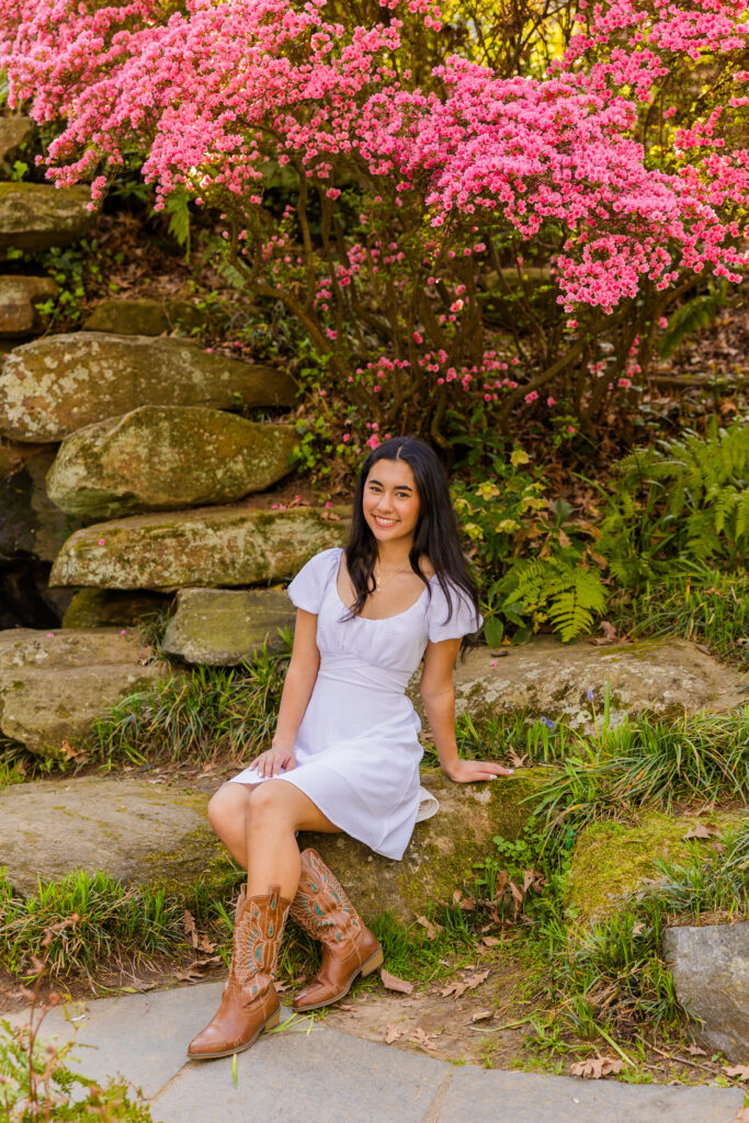 Senior sitting on rocks under pink flowers in Atlanta park wearing white dress