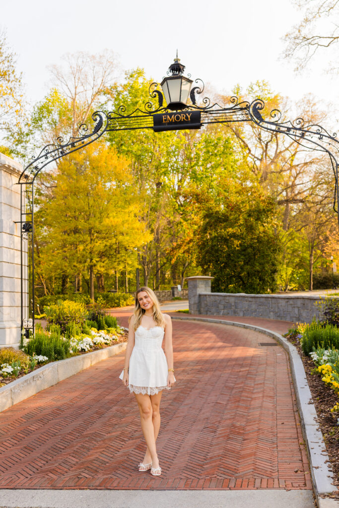 Colbie standing under Emory entrance arch with flowers wearing white dress graduation photos Atlanta