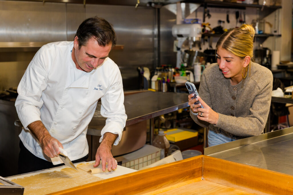 Chef cutting dough while daughter films content in kitchen