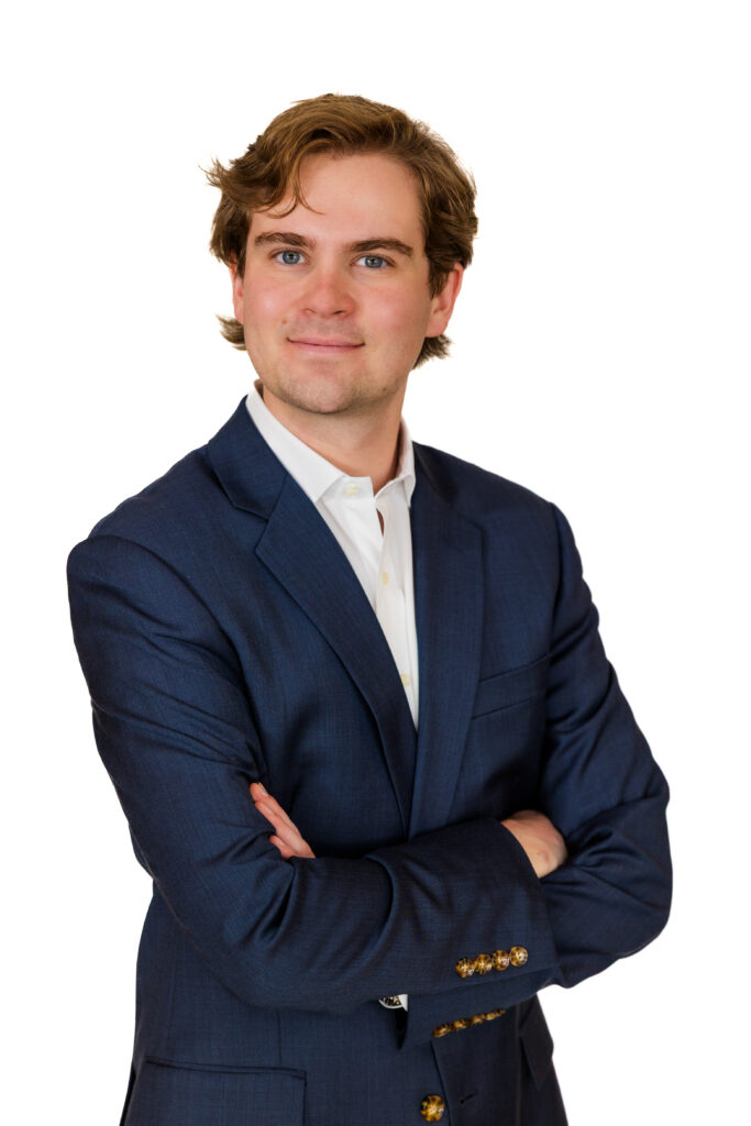 Professional corporate headshot of man in dark brown blazer, arms crossed, white backdrop.