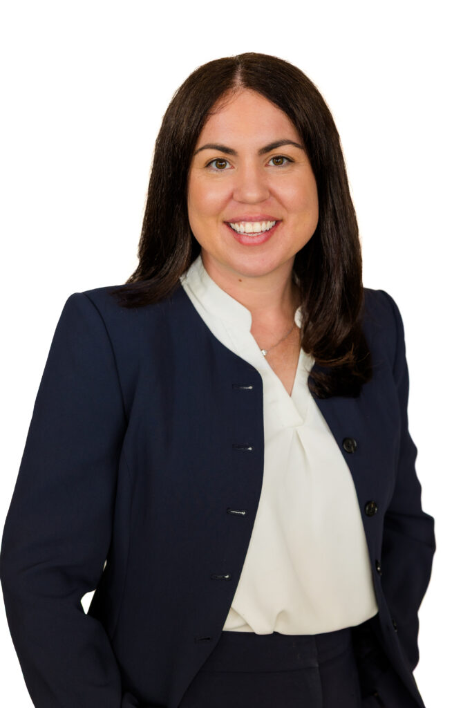 Professional business headshot of woman with long brown hair in blue blazer, hands in pockets, white background.