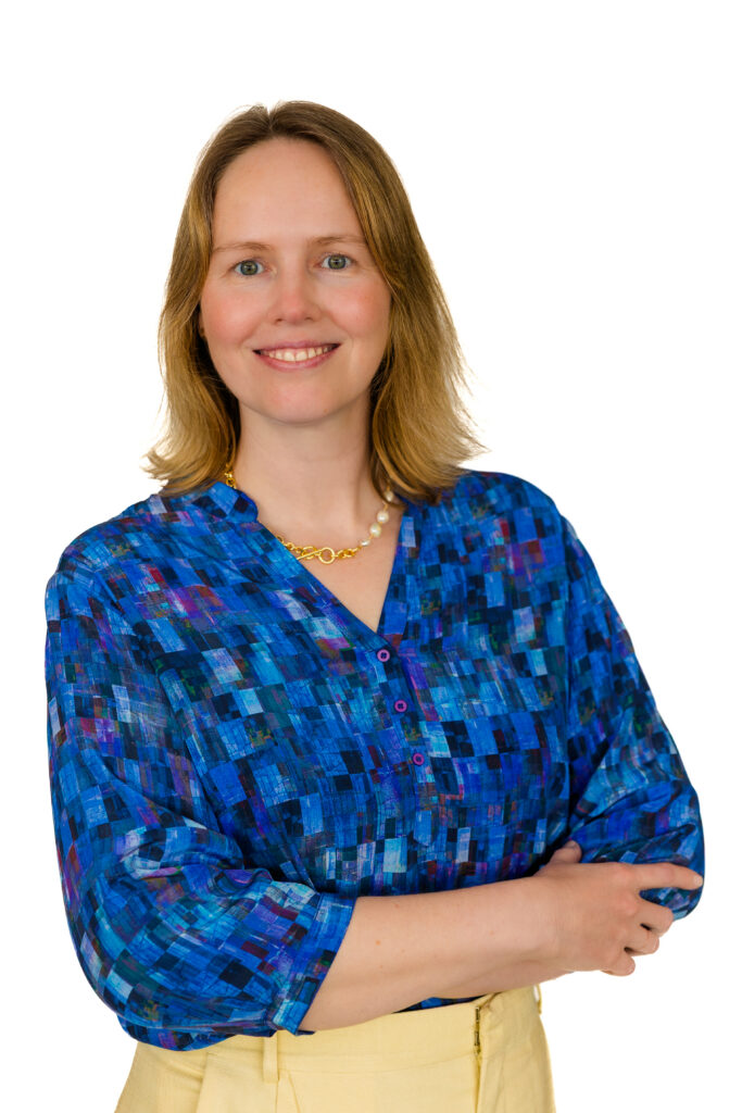 Professional headshot of woman wearing yellow pants and blue shirt, arms crossed, white backdrop.