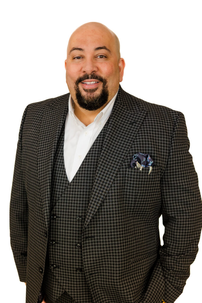 Corporate headshot of man in brown plaid suit, hands in pockets, angled pose, white background.
