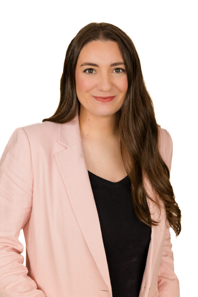 Corporate headshot of woman in pink jacket, hands in pockets, 45 degree angle, white background.
