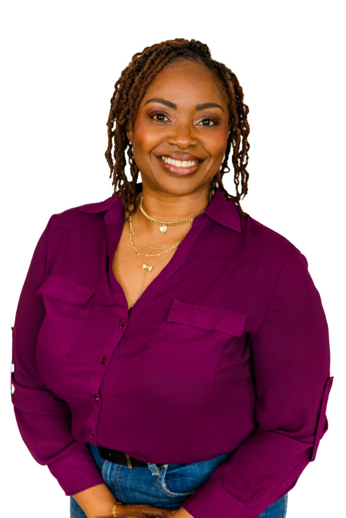 Professional corporate headshot of woman in purple shirt and jeans, hands together, white backdrop.