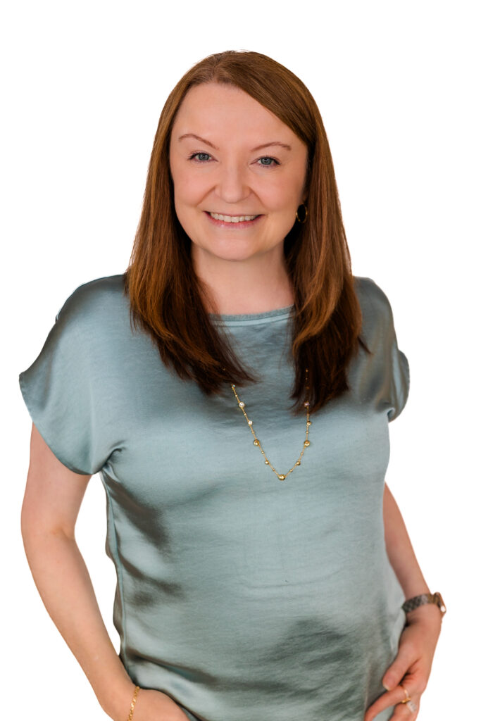 Professional business headshot of woman in light green silk top, hands in pockets, white backdrop.